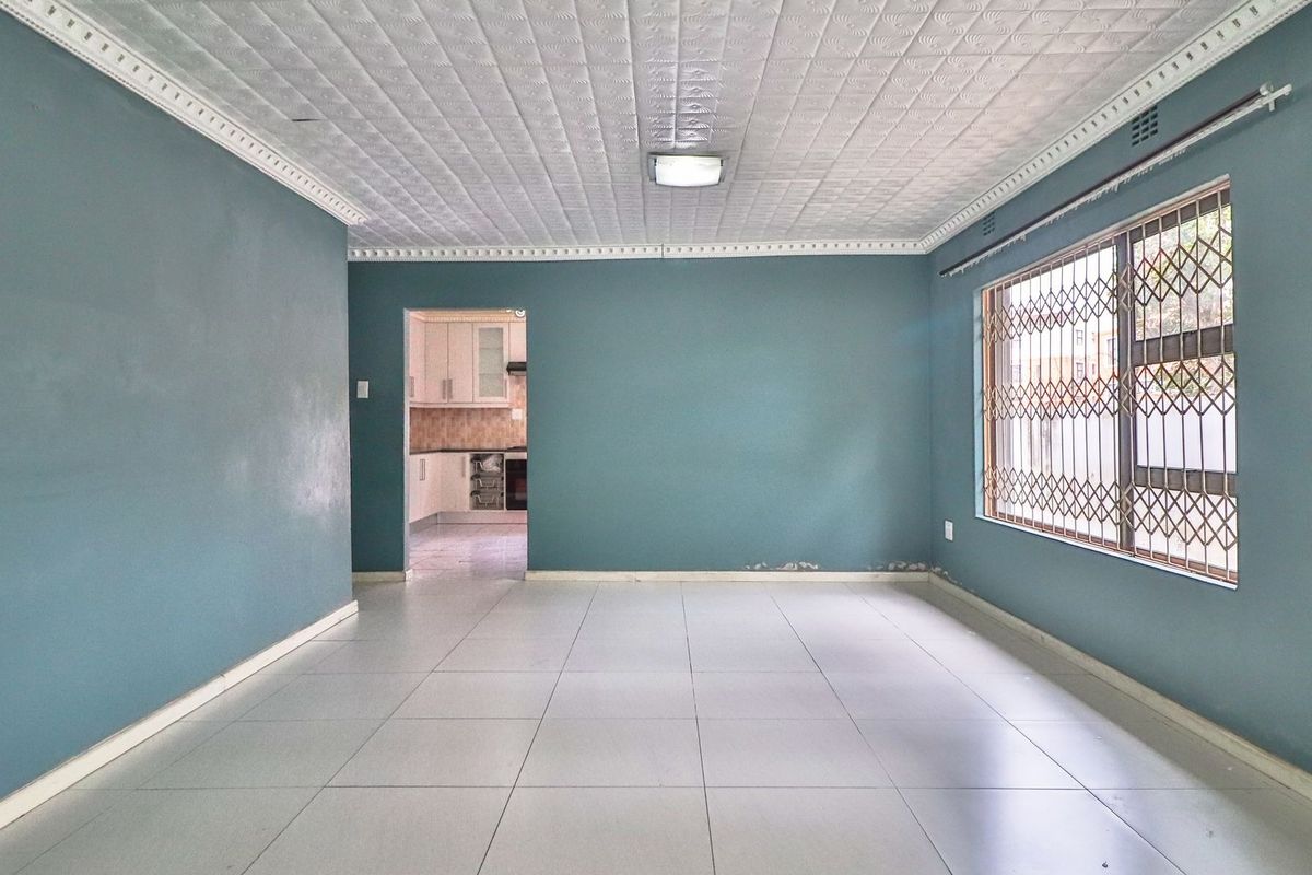 An empty living room featuring tiled floors, decorative ceiling tiles, and a window with a security gate, leading through to a kitchen area.
