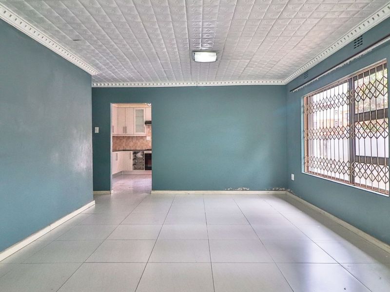 An empty living room featuring tiled floors, decorative ceiling tiles, and a window with a security gate, leading through to a kitchen area.