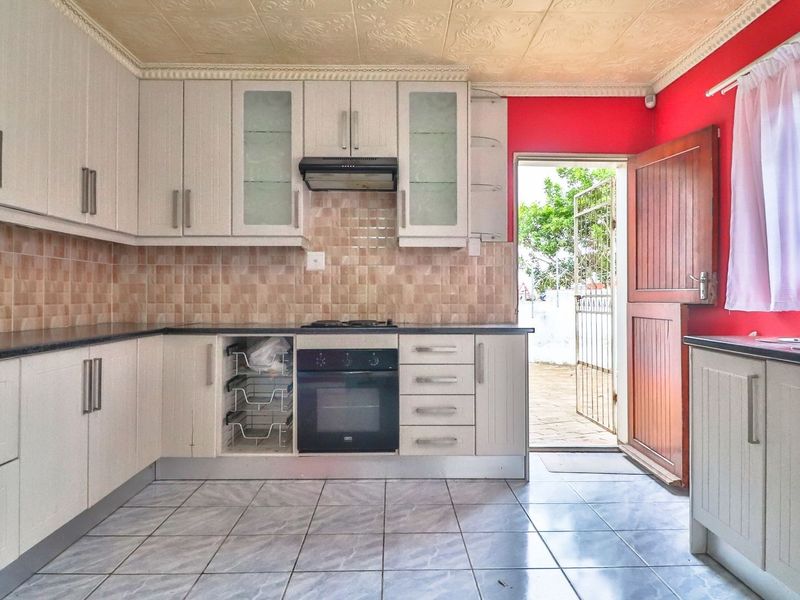 A functional kitchen featuring white built-in cabinetry, tiled backsplash, integrated oven and hob, and direct access to the exterior via a timber door.