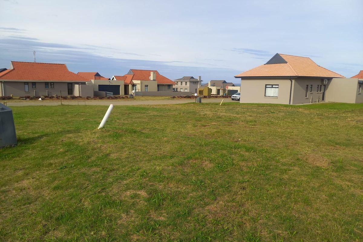 A vacant residential plot of land featuring a grass lawn with surrounding neighborhood houses visible in the background.