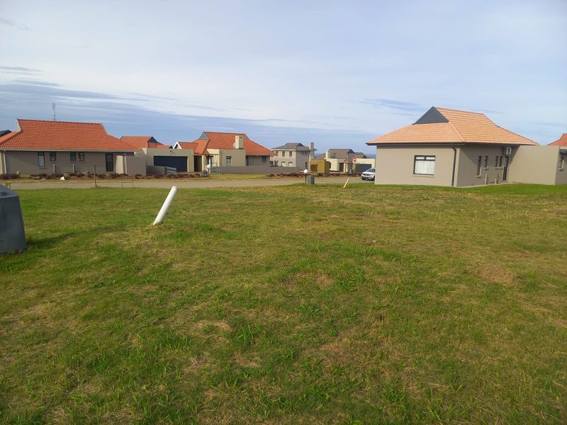 A vacant residential plot of land featuring a grass lawn with surrounding neighborhood houses visible in the background.