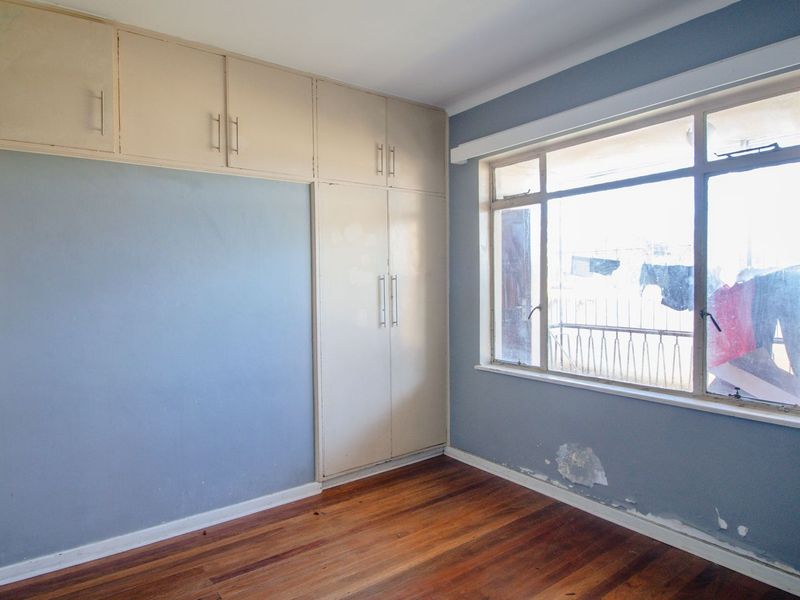 An empty bedroom featuring wooden flooring, built-in cupboards, and a large window. The walls show signs of wear and require maintenance.