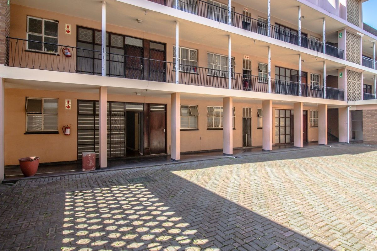 Exterior view of a multi-story apartment block featuring a paved courtyard, covered walkways, and balcony access with wrought iron railings.