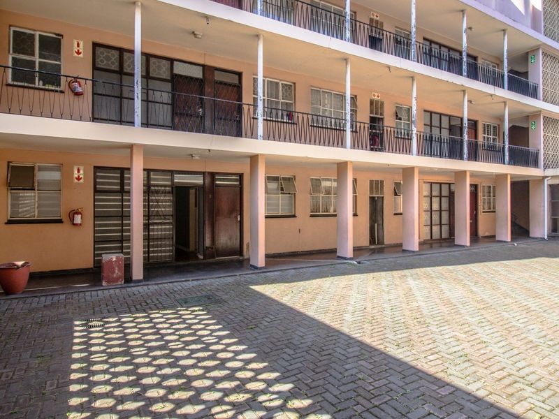 Exterior view of a multi-story apartment block featuring a paved courtyard, covered walkways, and balcony access with wrought iron railings.