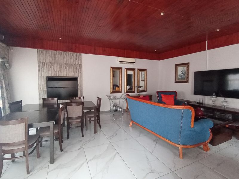 Spacious open-plan living and dining area featuring a built-in fireplace with stone cladding, polished porcelain floor tiles, and a warm timber ceiling.