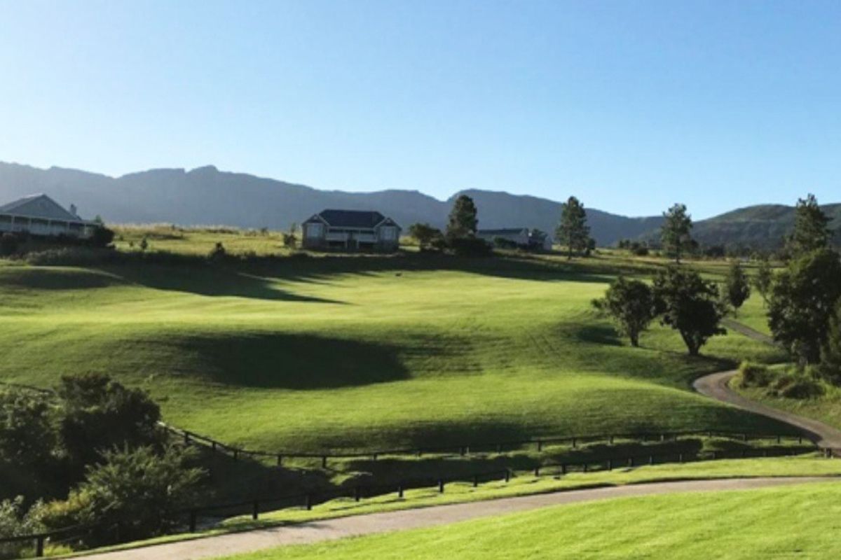 A scenic view of a rolling green landscape with established residential properties in the distance, featuring paved access roads and post and rail fencing.