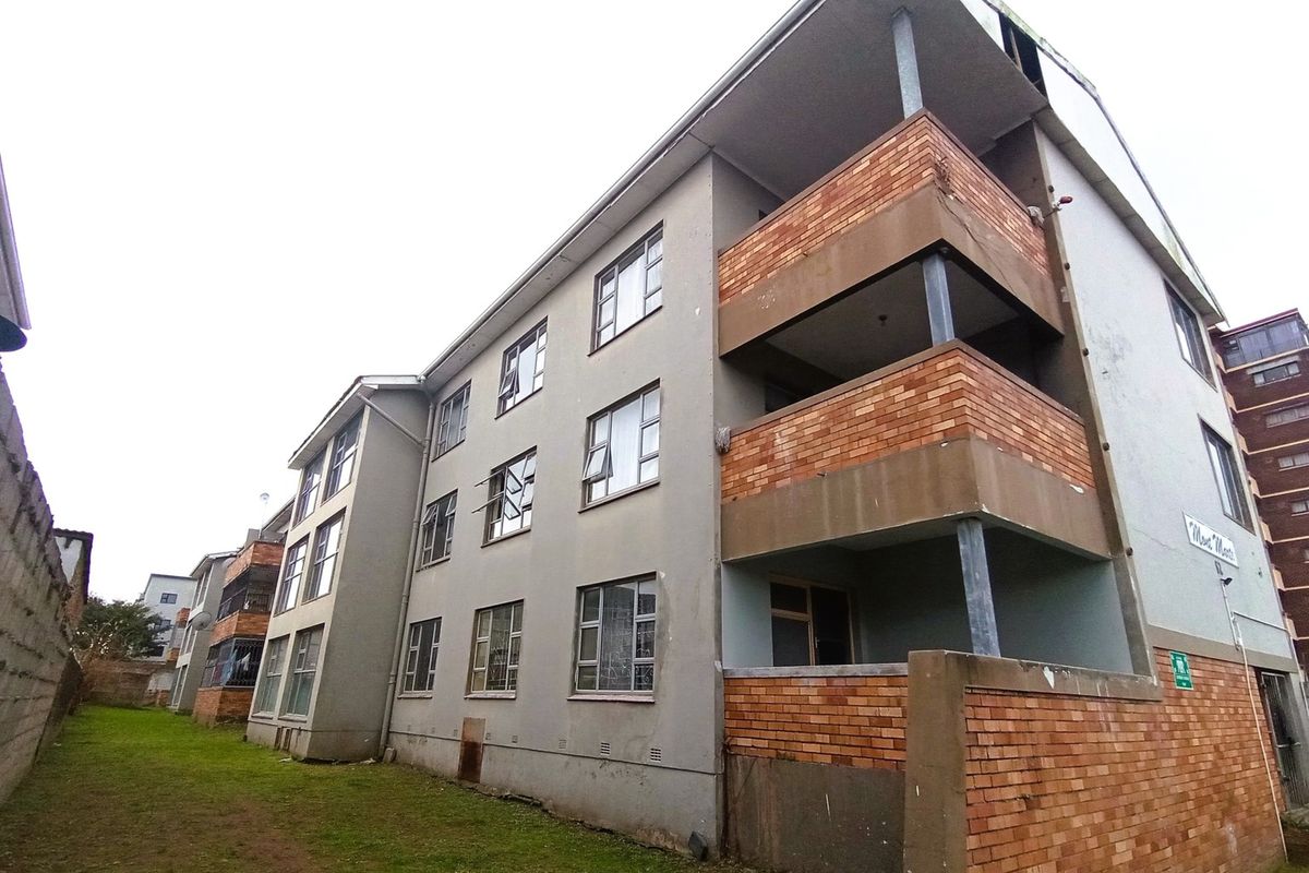 Rear view of a multi-story apartment building featuring brick balconies, metal-framed windows, and a shared grass area.