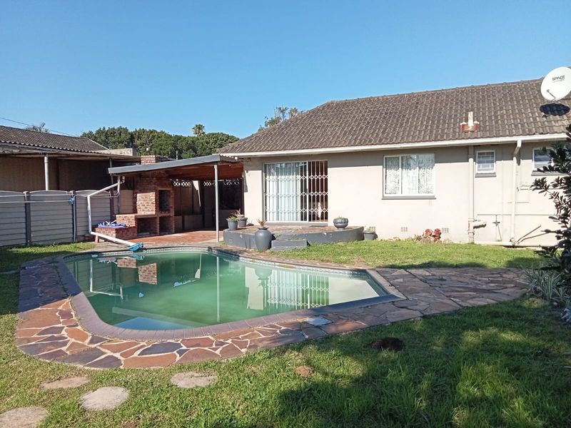 Rear exterior view featuring a swimming pool, stone-paved surround, and a covered patio area with a built-in brick braai.