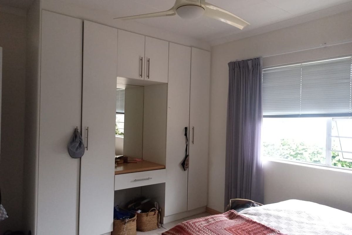 A well-maintained bedroom featuring extensive white built-in cupboards with an integrated vanity area, ceiling fan, and window blinds.