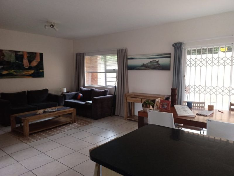 Open-plan living and dining area featuring tiled flooring, large windows with blinds, and a security gate leading to the exterior.
