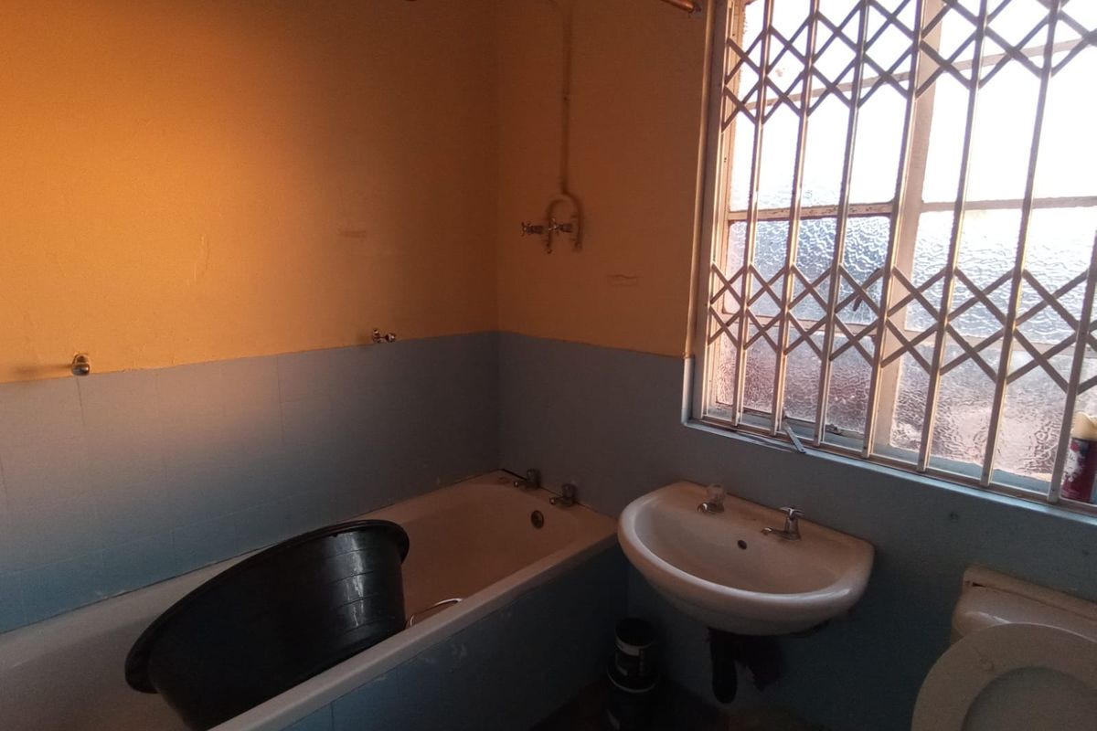 A dated bathroom featuring a bathtub, pedestal basin, and toilet, with security bars on the window and partially tiled walls.