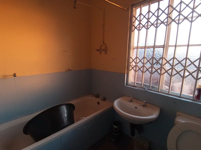 A dated bathroom featuring a bathtub, pedestal basin, and toilet, with security bars on the window and partially tiled walls.