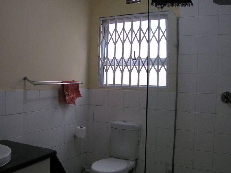 A functional bathroom featuring a shower, toilet, and vanity basin with tiled walls and a security gate on the window.