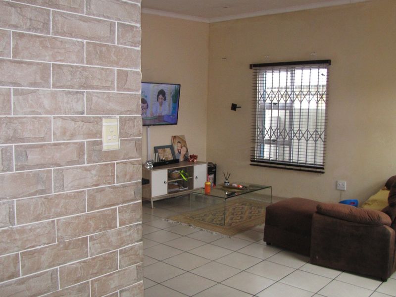 A living room featuring tiled flooring, a stone-cladding accent wall, and a window fitted with security bars and venetian blinds.