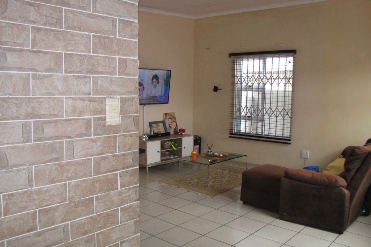 A living room featuring tiled flooring, a stone-cladding accent wall, and a window fitted with security bars and venetian blinds.