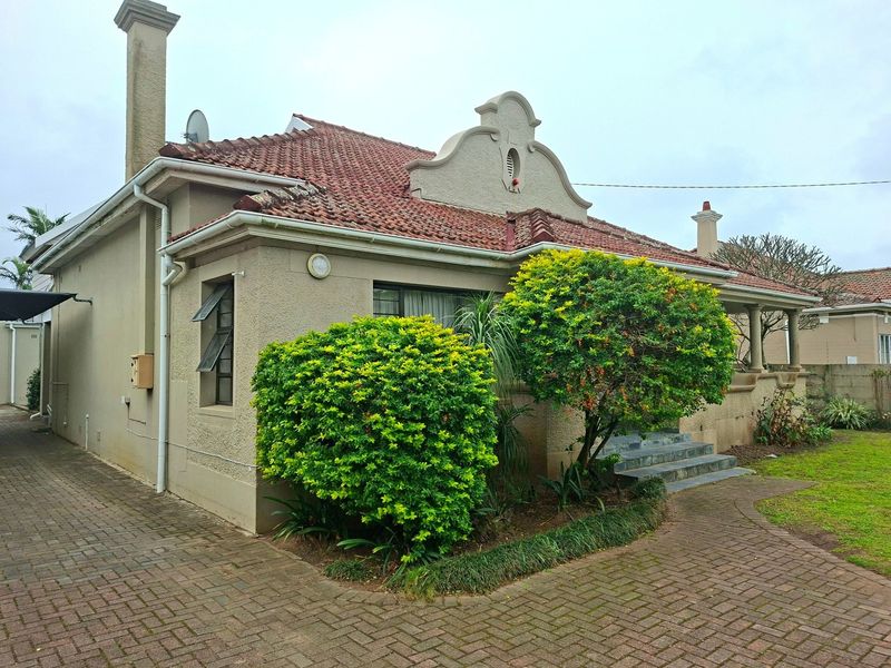 Front view of a charming house featuring a gabled roof, chimney, paved driveway, and well-maintained garden area.