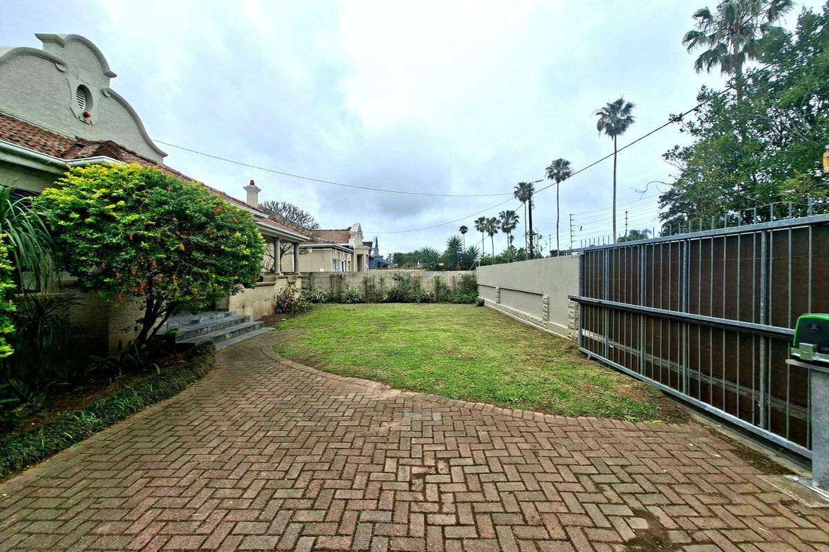 A spacious rear garden area featuring a paved driveway, lawn, and secure boundary wall with a sliding gate.