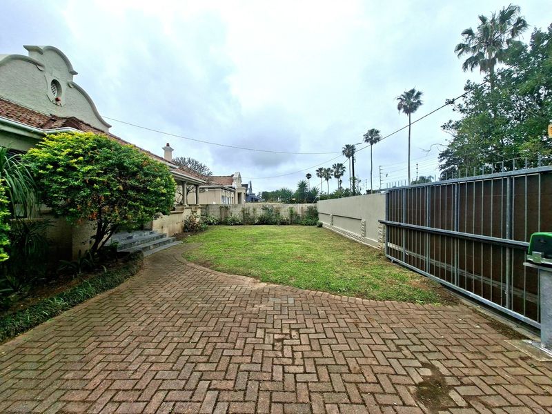 A spacious rear garden area featuring a paved driveway, lawn, and secure boundary wall with a sliding gate.