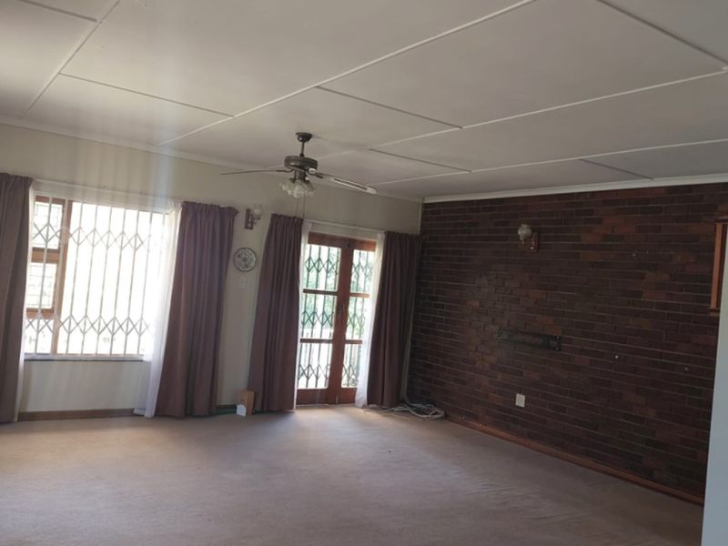 Spacious living room featuring an exposed brick wall, ceiling fan, and large windows with security gates, offering a blank canvas for renovation.