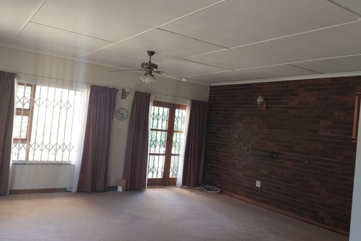 Spacious living room featuring an exposed brick wall, ceiling fan, and large windows with security gates, offering a blank canvas for renovation.