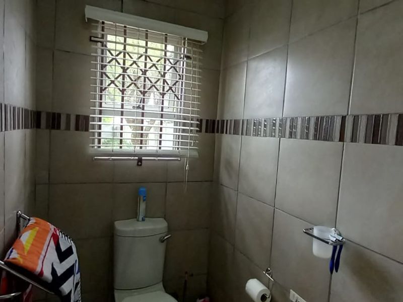 A functional bathroom featuring neutral ceramic wall tiles, a standard toilet, and a window with security bars and a venetian blind.