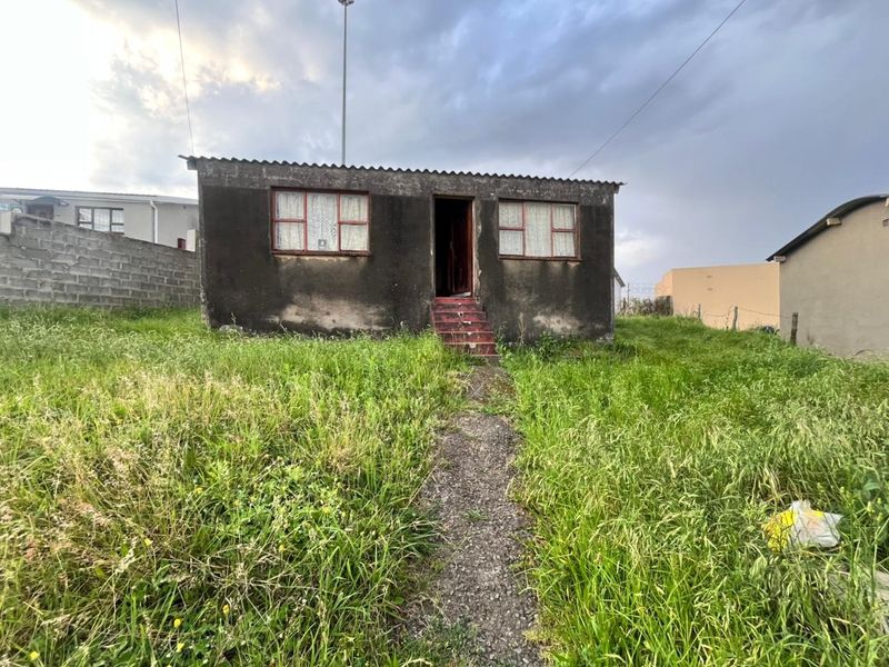 Front view of a detached house featuring a corrugated roof and metal window frames, set on a plot with overgrown grass and a concrete path.