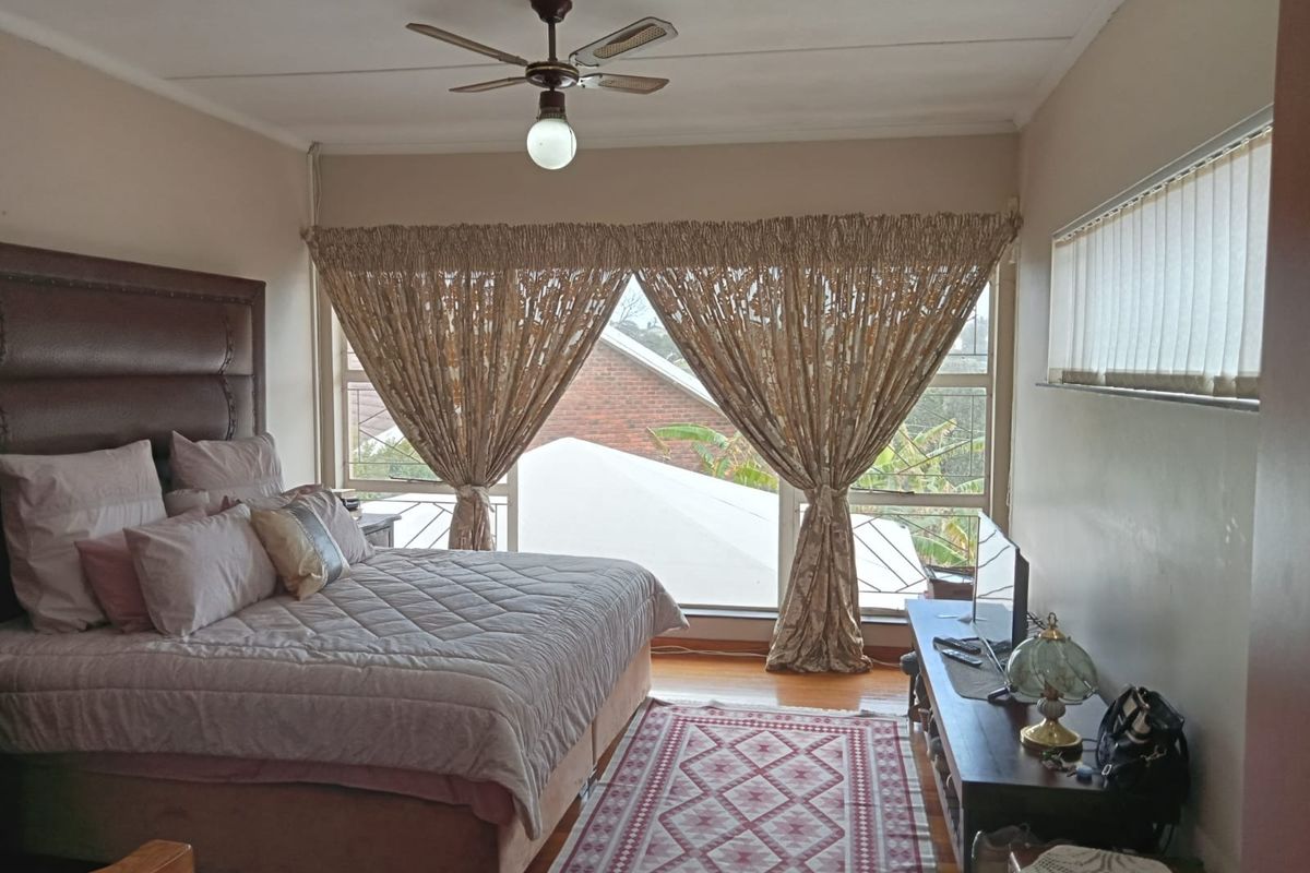 A spacious bedroom featuring hardwood flooring, a ceiling fan, and large windows with vertical blinds, offering natural light and a view of the surroundings.