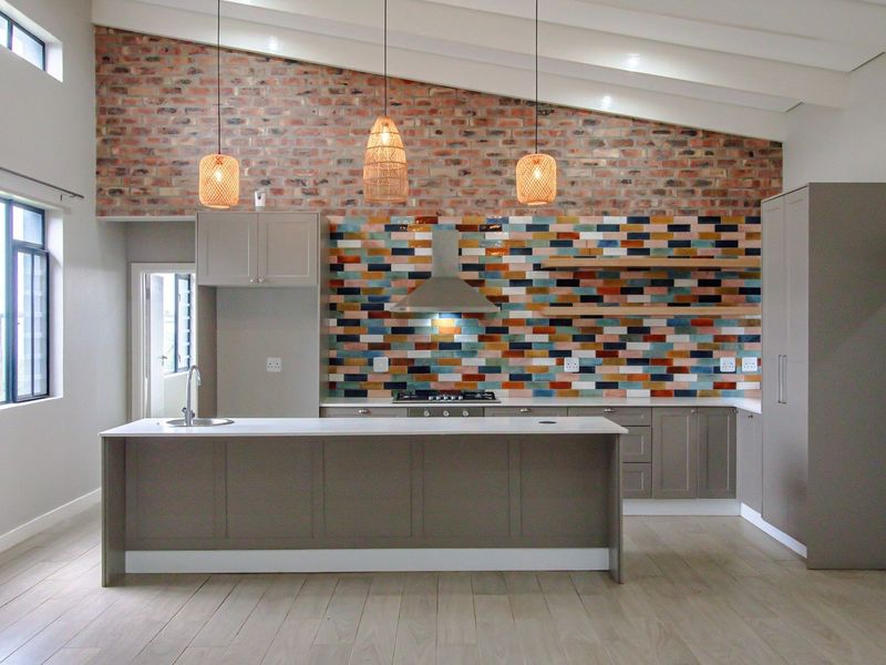 Modern kitchen featuring a central island, exposed brick feature wall, vibrant tiled splashback, and vaulted ceilings with contemporary cabinetry and high-quality finishes.