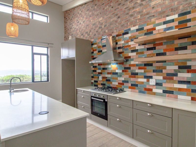 Modern kitchen featuring a large island, quartz countertops, built-in oven, gas hob, and a striking multi-colored tiled splashback against an exposed brick wall.