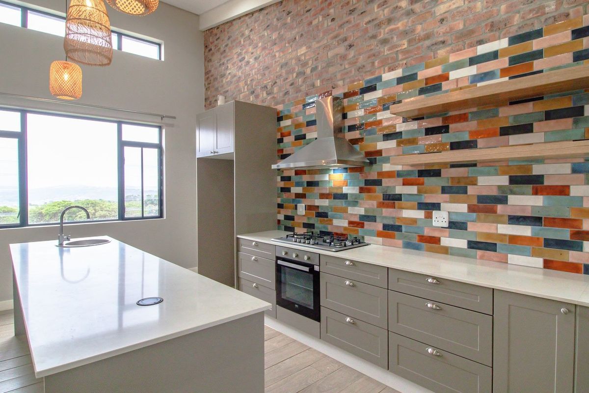 Modern kitchen featuring a large island, quartz countertops, built-in oven, gas hob, and a striking multi-colored tiled splashback against an exposed brick wall.