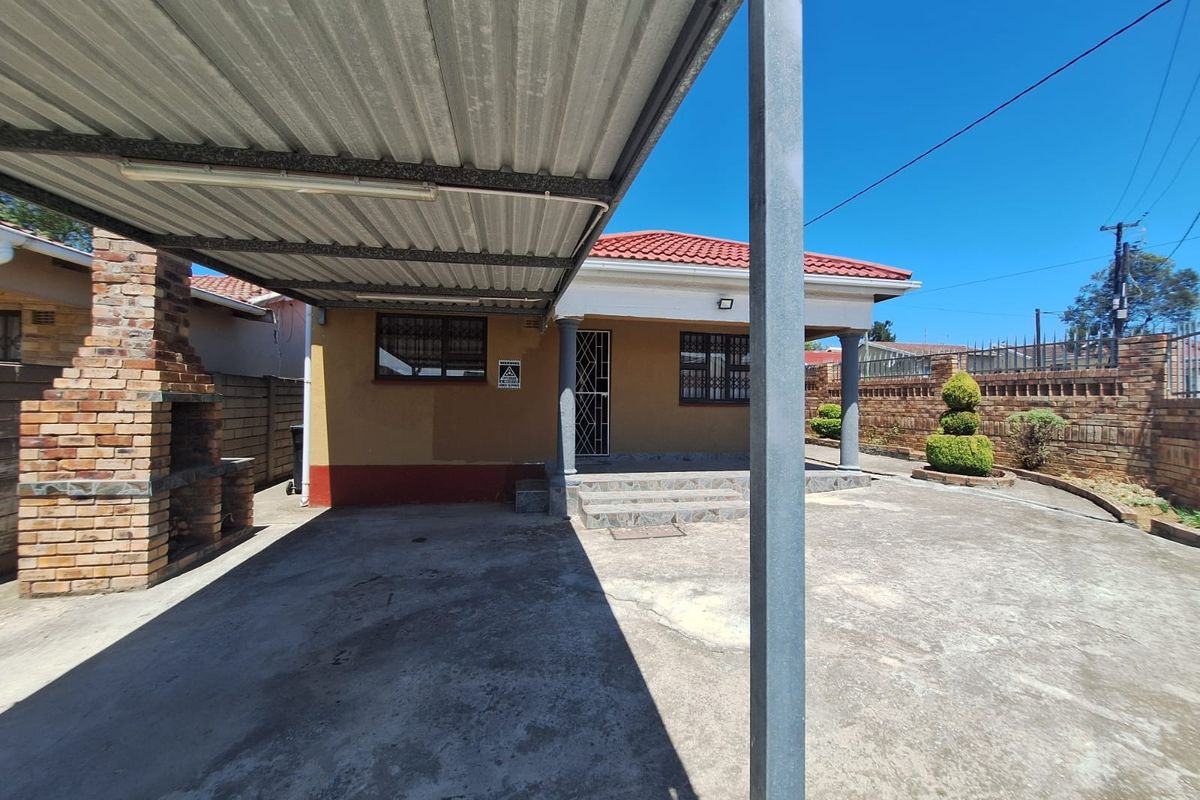 Rear exterior view featuring a covered carport, a brick built-in braai, and a paved driveway leading to the main house entrance.