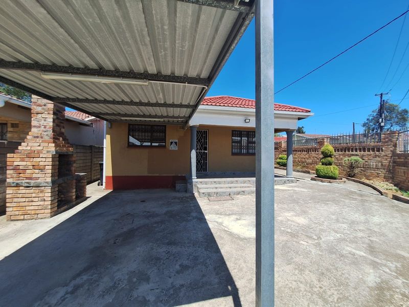 Rear exterior view featuring a covered carport, a brick built-in braai, and a paved driveway leading to the main house entrance.