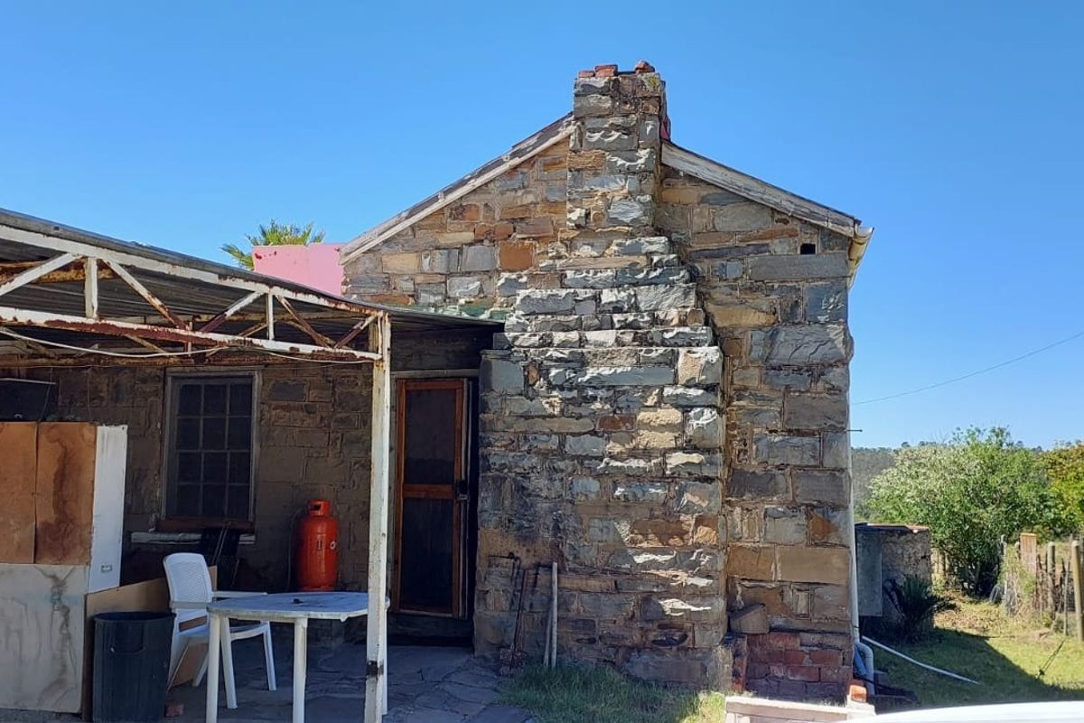 A rustic stone farmhouse exterior featuring a prominent stone chimney and an attached metal-framed carport area set within a rural landscape.