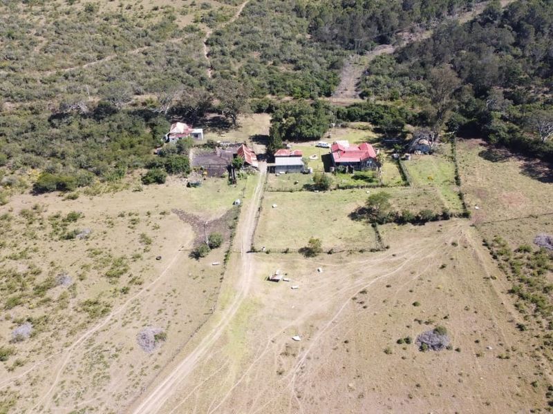 Aerial view of a rural farm property featuring multiple outbuildings, fenced paddocks, and an unpaved access road set within a natural landscape.