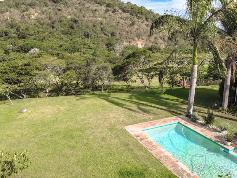 An elevated view of a swimming pool with brick paving, set within a spacious green garden against a scenic hillside backdrop.