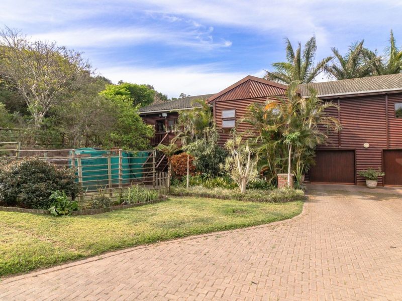 Front view of a timber-clad farm property featuring a brick-paved driveway, double garage, and established garden with water storage tanks.