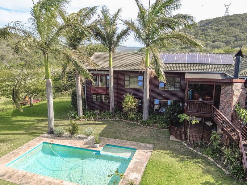 Aerial view of a timber-clad farmhouse featuring a swimming pool, raised wooden deck, and roof-mounted solar panels set against a scenic rural landscape.