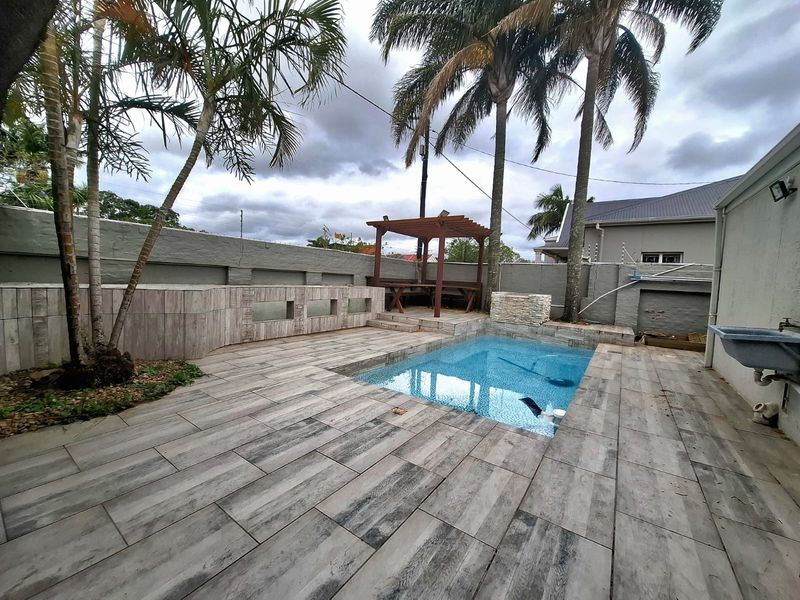Outdoor pool area featuring a tiled deck, wooden pergola, perimeter wall with electric fencing, and a wall-mounted utility sink.