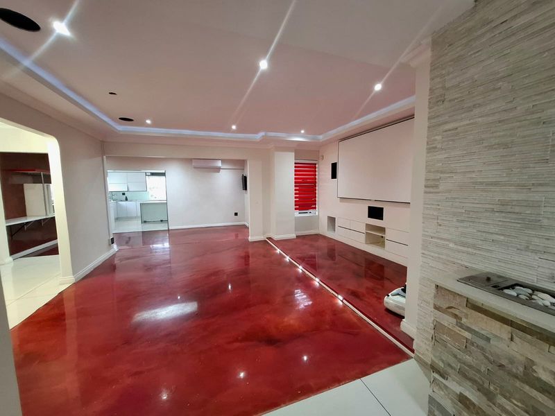 Spacious open-plan living area featuring high-gloss red epoxy flooring, recessed ceiling lighting, a stone feature wall with built-in braai, and a custom media unit.