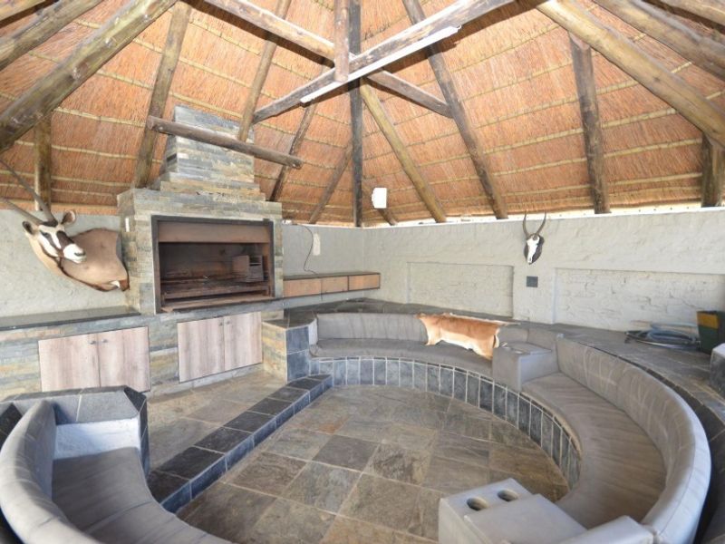 An outdoor entertainment lapa featuring a thatched roof, exposed timber rafters, built-in braai with stone cladding, and integrated circular stone seating.