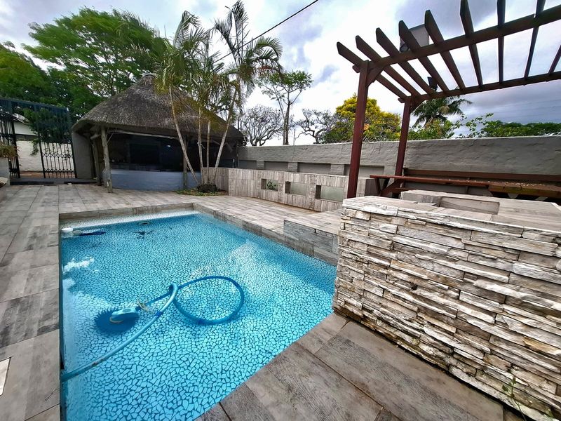 Outdoor entertainment area featuring a swimming pool, stone-clad built-in braai, wooden pergola, and a thatched lapa in the background.
