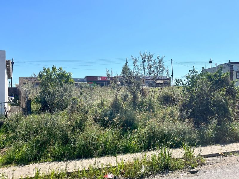 A vacant residential plot featuring overgrown vegetation and a paved sidewalk, situated in a developed area with nearby commercial infrastructure.