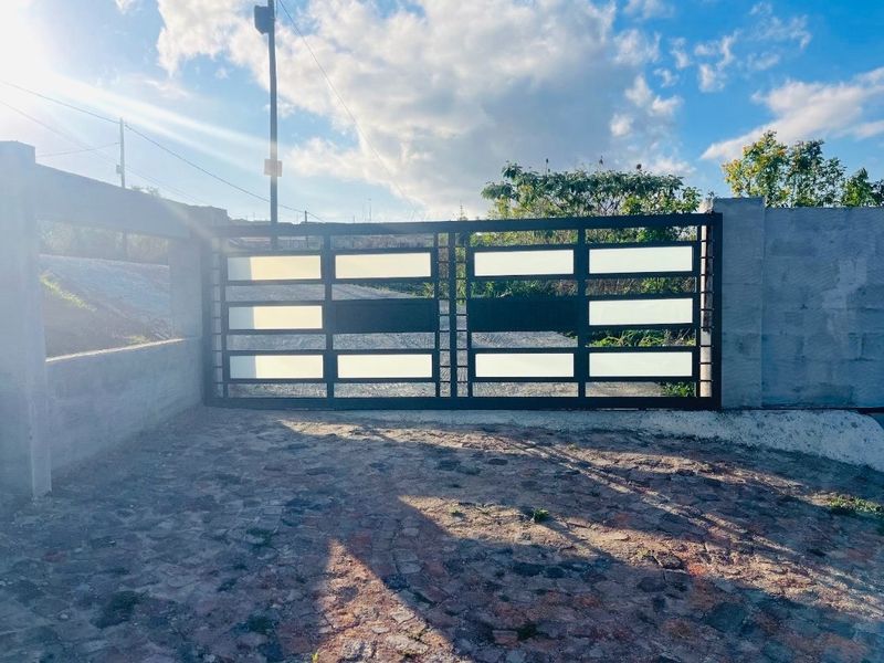 A modern metal driveway gate set within a concrete boundary wall, featuring a cobblestone driveway approach.