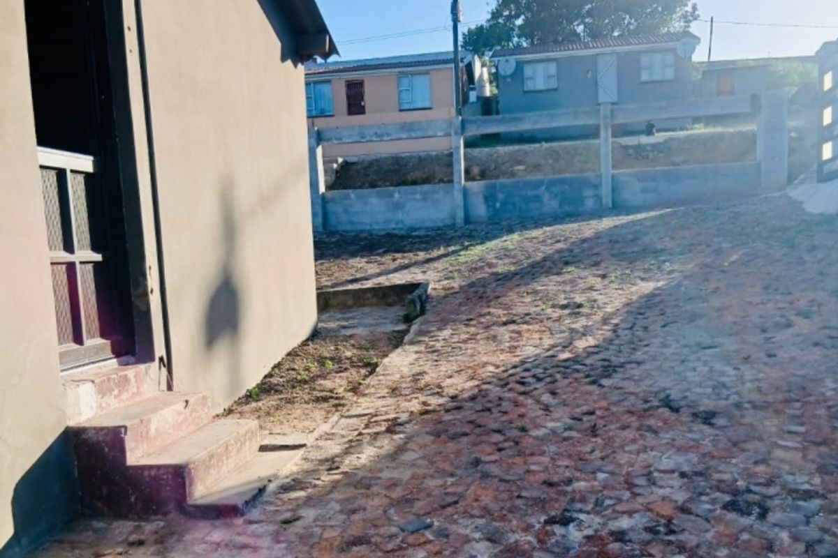 Rear exterior view showing a cobblestone paved area, concrete boundary wall, and access steps leading to the house entrance.