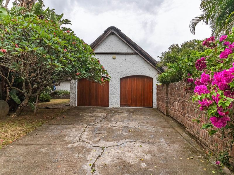 A detached double garage featuring wooden doors and a thatched roof, situated at the end of a concrete driveway with surrounding garden landscaping.