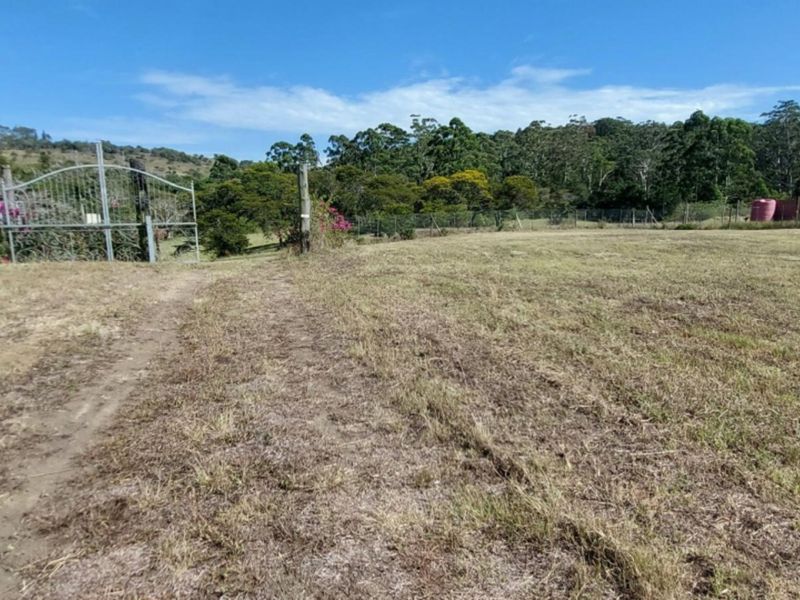 A view of a vacant residential plot featuring a metal entrance gate, perimeter wire fencing, and a water storage tank in the background.