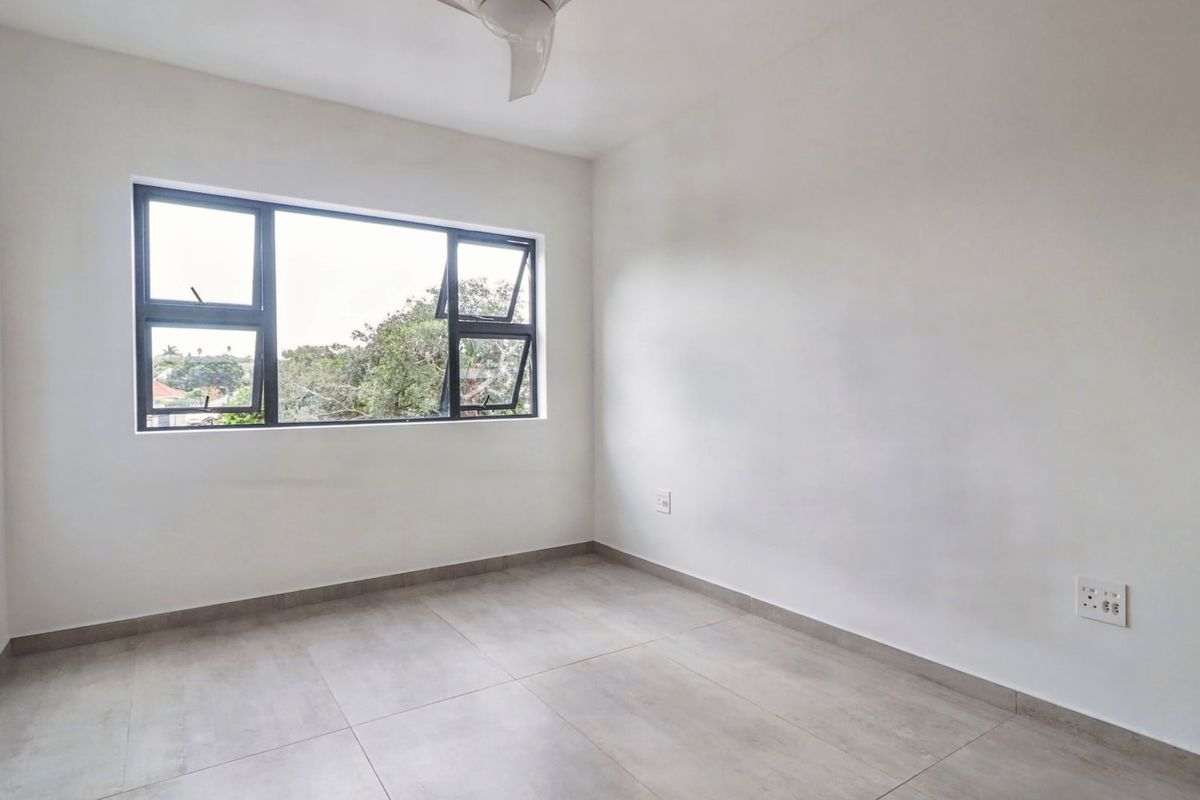 A bright, empty bedroom featuring neutral tiled flooring, a ceiling fan, and large aluminium-framed windows providing natural light.