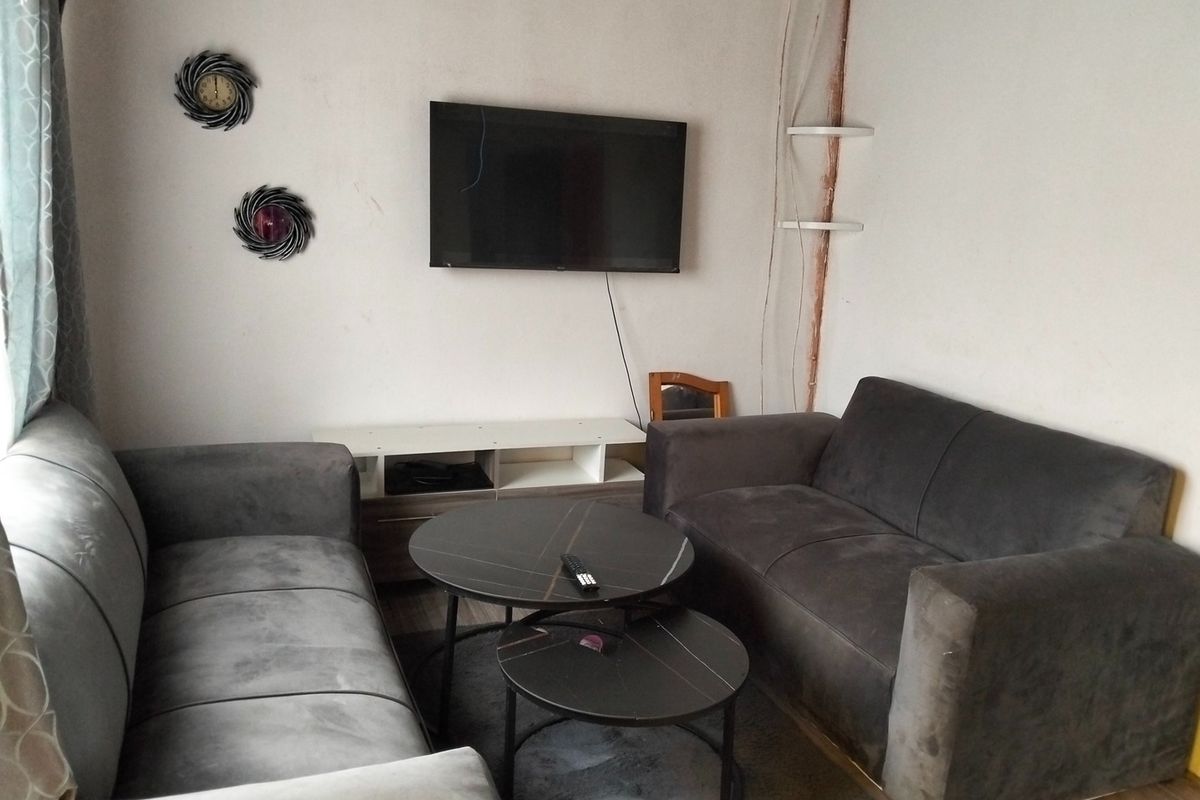 A compact living room featuring laminate flooring and a wall-mounted television, currently showing signs of wear including exposed wiring and damaged wall surfaces.