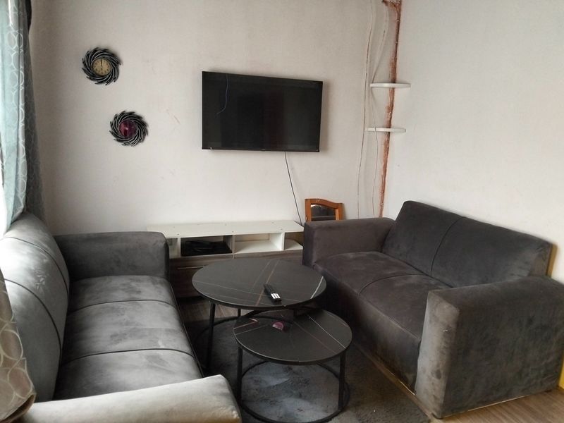 A compact living room featuring laminate flooring and a wall-mounted television, currently showing signs of wear including exposed wiring and damaged wall surfaces.