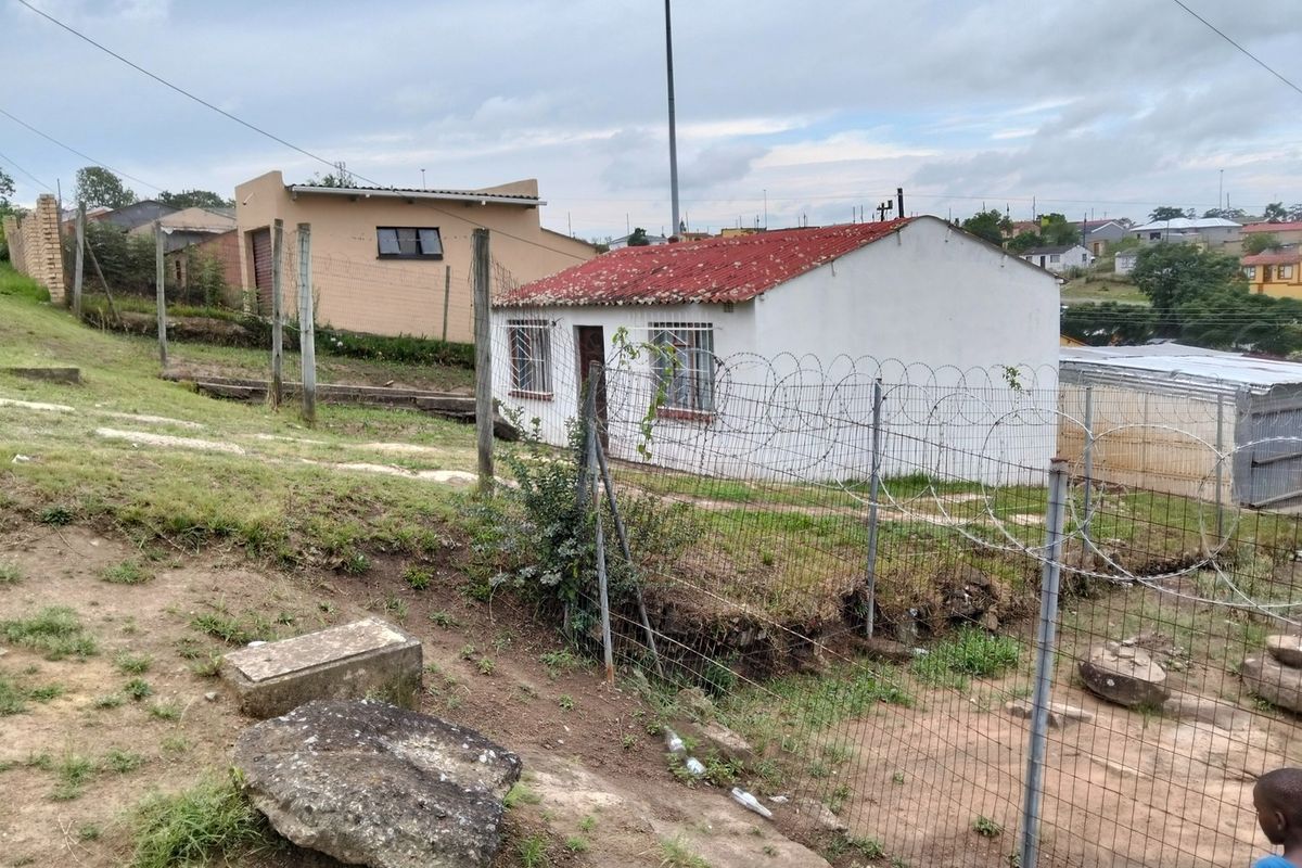 Exterior view of a small residential structure featuring a corrugated metal roof and security fencing in a suburban setting.